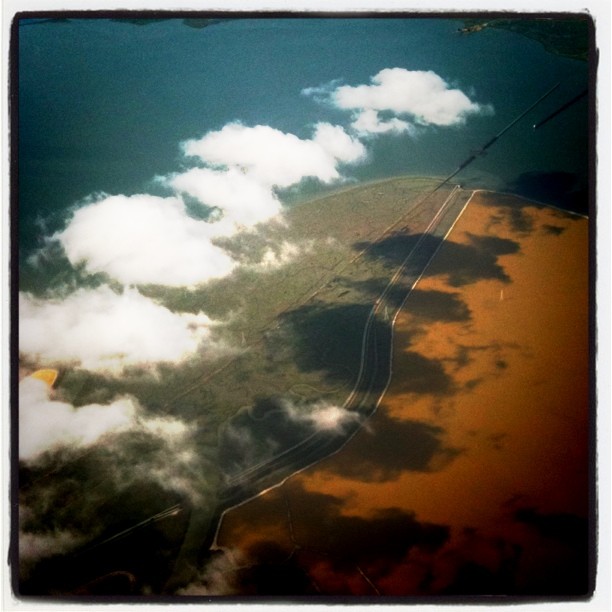 airplane window photo of clouds casting shadows over teal, gold, and brown sections of land