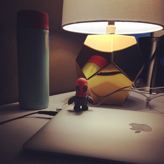 dimly lit desk with Zoidberg toy standing on a closed Macbook next to a geometric, gold lamp