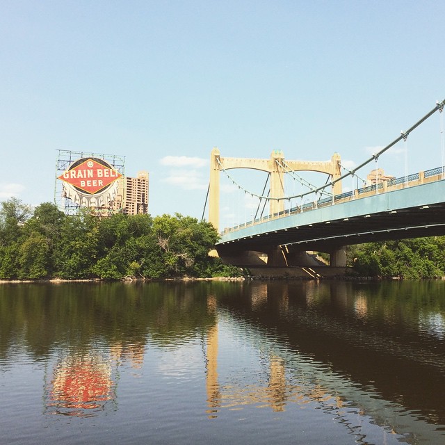 river with blue steel bridge; a Grain Belt Beer sign is across the way above some bushes