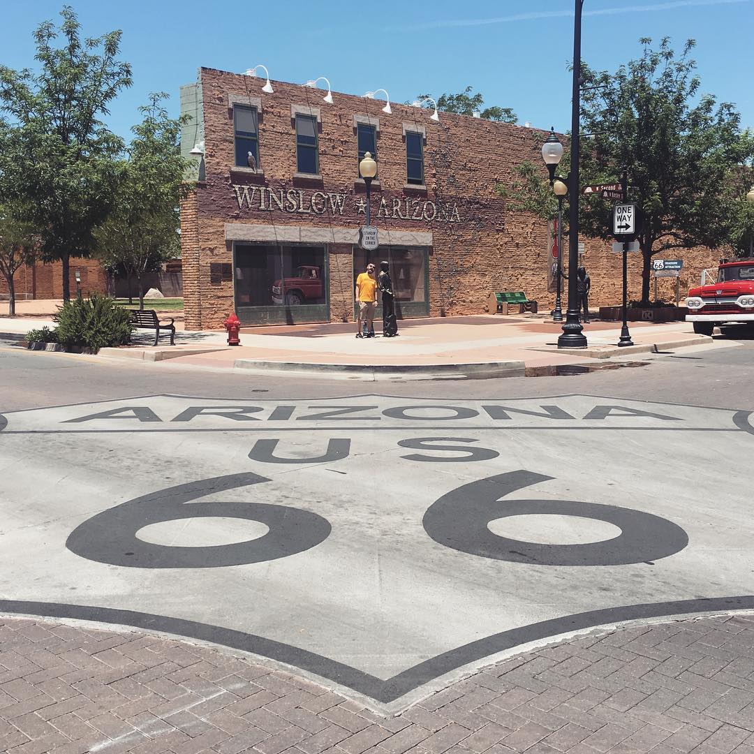 Clay standing on a street corner. Route 66 sign painted big in the intersection. A brick wall behind is painted with Winslow, Arizona